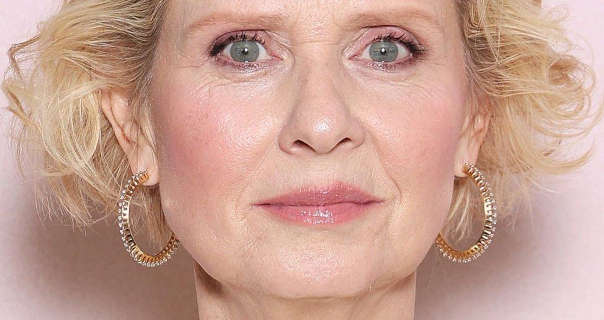 Cynthia Nixon wears her blonde hair cut short and in frizzy curls. She stands in front of a pale pink wall. Her makeup is a dark pink lipstick, blush and clumpy mascara. She wears a pair of large metal gold hoop earrings.