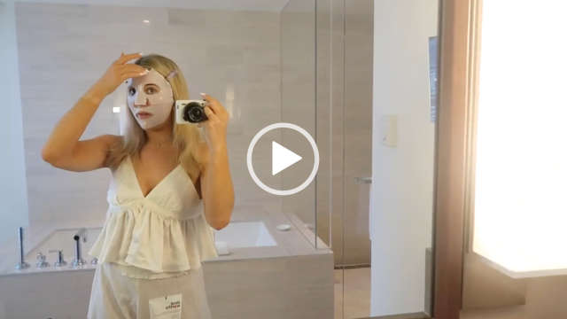 Em Sheldon adjusts a sheet face mask she has on. She looks at herself in a large mirror in her hotel room in New York. She stands in the cream tile bathroom next to a large light and in front of the bath. Em is seen with her blonde hair down and straight. She wears a cropped white camisole and a pair of slightly sheer off-white shorts.