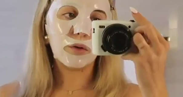 Em Sheldon records herself using a small white and black camera. She stands in a shiny off-white tile bathroom. Em is seen with her blonde hair loose and straight. She wears a sheet face mask with a white sleep top, gold earrings and a thin gold necklace.