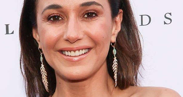 Emmanuelle Chriqui wears her black hair naturally loose and pushed back over her shoulders. She appears in front of a dark white wall with black text on it. Emmanuelle is seen in a light pink gloss lipstick, black mascara and a pair of hanging gold feather earrings.