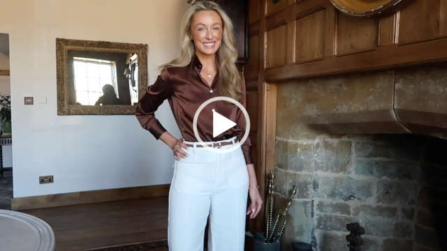 Josie Irons smiles as she looks towards the camera. She stands next to a large stone and wood panel open fireplace in a reception area of her countryside house. Josie has her blonde hair loose and in soft waves. She wears a pair of white trousers with a brown blouse from LilySilk.