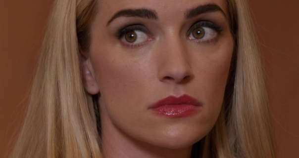 Brianne Howey looks to one side with a serious expression. She wears her thick blonde hair straight and down. Brianne appears in a dark red glossy lipstick and black eye shadow. She sits in front of a dark terracotta colour wall.
