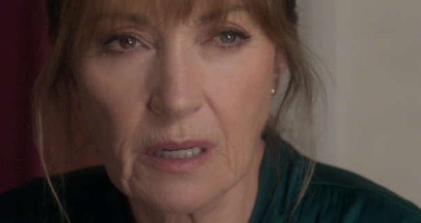 Jane Seymour appears with her hair tied up and with a few strands pulled down to frame her face. She wears a dark green slightly shiny top. Jane sits in front of an off-white wall and a wooden kitchen cabinet. She wears a pair of small stud earrings and a pink lipstick.
