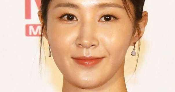 Kwon Yu-ri stands in front of an off white and pale red logo wall. She wears her black hair tied up. She is seen in a glossy lipstick, white and gold drop earrings and black mascara.