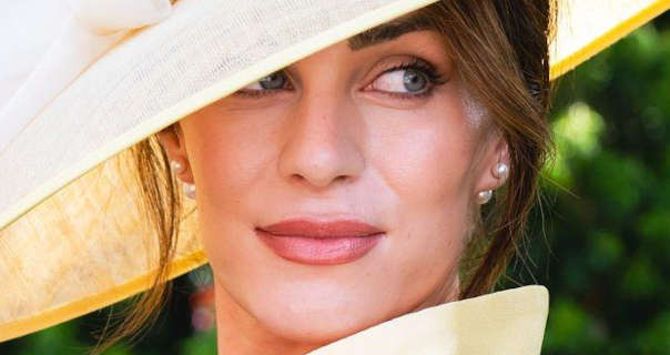 Lydia Elise Millen is seen standing in front of green leafy plants in dappled sunlight. She wears a wide brim yellow hat with a structured yellow dress. Lydia is seen in pearl earrings, a glossy dark pink lipstick and black mascara.