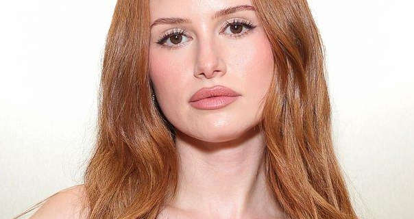 Madelaine Petsch appears in front of a white to silver gradient wall. She wears her long red hair is loose and in natural waves. Madelaine is seen in a dark pink matt lipstick, pink blush and black mascara.