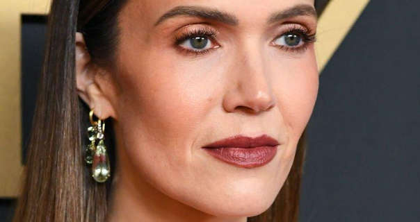 Mandy Moore gazes to one side. She stands in front of a dark grey wall that has gold text on it. Mandy is seen with her long brown hair straight and loose. She wears a pair of large gold and glass drop earrings, black mascara, contouring foundation and a deep red lipstick.
