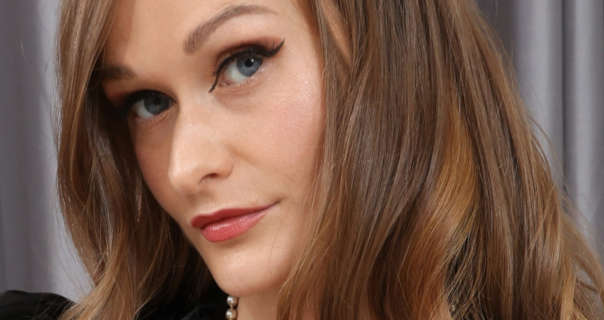 Honour May is seen in front of a pair of light grey drawn curtains. Her light brown hair has soft highlights and is loose and down. She wears black winged eyeliner with a dark red gloss lipstick and black mascara. Honour appears in a black top and a pearl necklace.