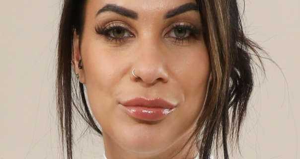 Roxy Mendez stands in front of a pale beige wall. She wears has blonde highlights in her black hair that is tied up and back in a bun. Roxy appears in a very glossy pink lipstick, a single silver nose ring, stud earrings and heavy black mascara.