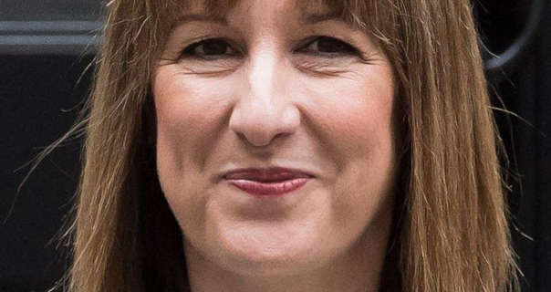 Rachel Reeves smiles while standing in front of the closed black door of 10 Downing Street. Her light brown hair is straight and down. Rachel wears black eyeliner and a dark red lipstick.