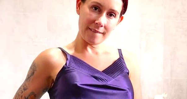 Belle O'Hara bits her bottom lip. She stands in front of a tiled bathroom wall. Belle has her black and red hair tied up in a bun. She is seen in a dark purple slip and no makeup.