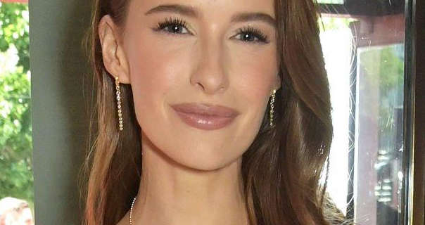 Victoria Magrath stands in front of a large glass window on a sunny day. She wears her light brown hair loose and in soft waves. Victoria is seen in a dark pink gloss lipstick, mascara and a pair of thin hanging gemstone earrings.