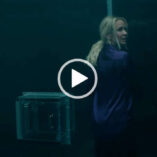 Anna Camp is seen in a large glass and metal box room next to a transfer hatch. She wears her long blonde hair down and tied back in a ponytail. Anna looks to one side. She wears a dark purple satin top with black trousers.