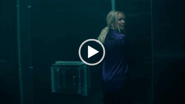 Anna Camp is seen in a large glass and metal box room next to a transfer hatch. She wears her long blonde hair down and tied back in a ponytail. Anna looks to one side. She wears a dark purple satin top with black trousers.