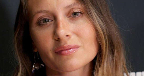 Aly Michalka is seen in front of a dark grey poster wall that has white block writing on it. She wears her long light brown hair loose and with soft highlights. Aly stars in a dark pink gloss lipstick, a little mascara and a pair of hanging metal earrings.