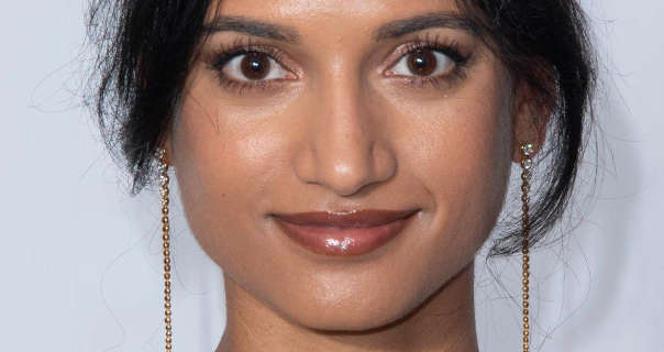 Amita Suman stands with her black hair tied up and back. She wears a pair of long thin dangling small bead earrings with a dark mauve gloss lipstick and curled eyelashes. She is seen in smiling in front of an off-white wall.