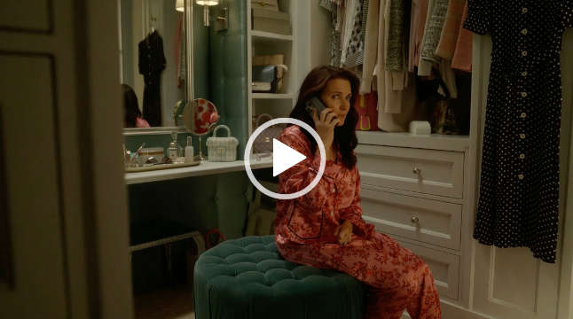 Kristin Davis sits on a large dark teal footstool in a white wood dressing room. She talks on a mobile phone and glances upwards. Kristin is seen in a pair of salmon and dark pink long sleeve pyjamas. She has her hair loose and in soft waves.