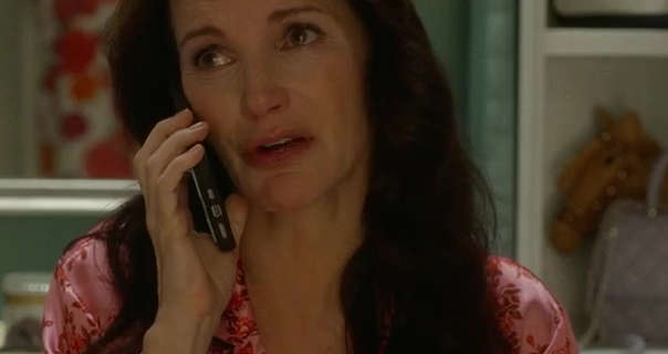 Kristin Davis holds a mobile phone up to one ear. She sits in a white wood walk in dressing gown. Kristin wears her thick black hair naturally loose. She is seen in a red lipstick, pink pyjamas and black mascara.