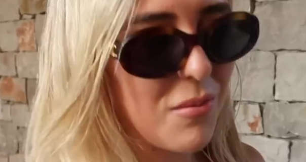 Em Sheldon walks past a rustic brick wall on holiday in Greece. She wears a pair of large oval frame sunglasses. Em is seen with her thick blonde hair loose and straightened. She wears a dark pink lipstick.