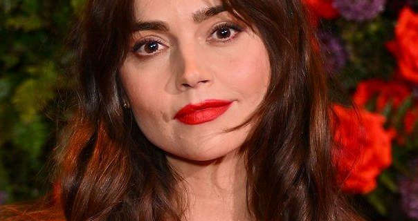 Jenna Coleman is seen in front of a red and purple rose with green leaf display wall. She wears her long thick hair loose and in soft waves. Jenna appears in black mascara, light blue and a dark red matt lipstick.