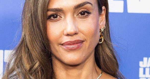 Jessica Alba is seen in front of a dark blue wall that has white writing on it in a chunky font. Jessica has her brown hair in waves and with highlights. She wears a dark red gloss lipstick, black mascara, a thin gold necklace and several gold hoop earrings.