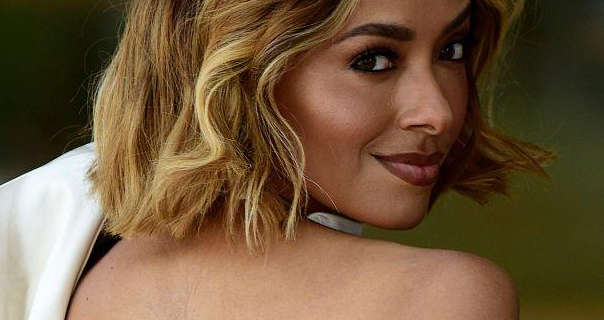 Kat Graham looks over her bare back and into the camera as she stands outdoors. She wears her hair cut short and coloured blonde. Kat is seen in a dark red lipstick, heavy eye shadow and black mascara.