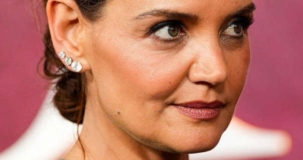 Katie Holmes wears a dark red sheeny lipstick and medium foundation. She has her hair tied back in a messy bun. Katie appears in front of a faded maroon wall with a large white letter on it. She wears three diamond earrings in increasing size.