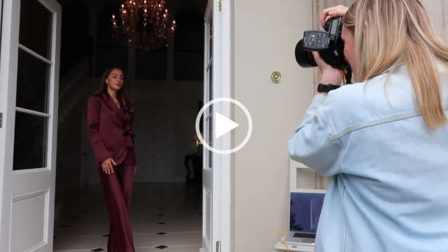 Lydia Elise Millen stands in a white wood frame doorway next to the open glass doors and with a lit chandelier behind her. She wears a silk burgundy suit. Lydia appears with her hair loose and pushed to one side. She is having her picture taken during a fashion campaign shoot.