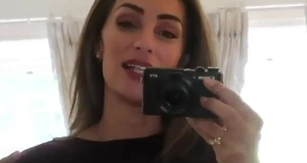 Lydia Elise Millen holds a Canon vlogging camera in one hand as she records herself through a mirror. She stands in front of a row of large windows in a cream bedroom in a manor house. She appears with her brown hair loose and straight. Lydia wears a burgundy cashmere jumper with a maroon lipstick and gold jewellery.
