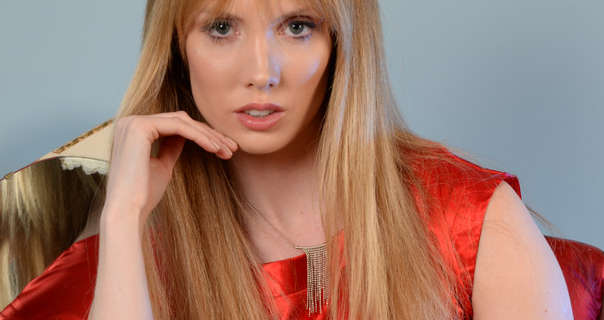 Elle sits in front of a dusty blue wall and a dressing table mirror. She wears her long blonde hair loose and with a fringe. Elle is seen in a sleeveless red dress, pink lipstick and clumpy black mascara.