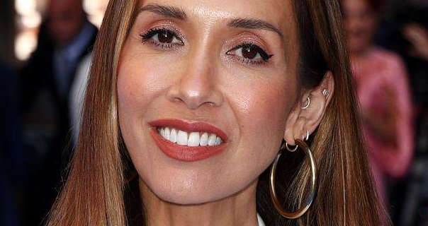 Myleene Klass is seen with her light blonde hair loose, down and with blonde streak highlights. She wears a dark red matt lipstick with a large gold hoop, stud and small circle earrings. Myleene stands in front of guest, interviewers and organisers at a public event.
