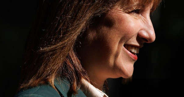 Rachel Reeves is seen in side profile against a black backdrop. She wears her shoulder length light brown hair loose. Rachel is seen in a dark forest green blazer, a pale gold blouse and red lipstick.
