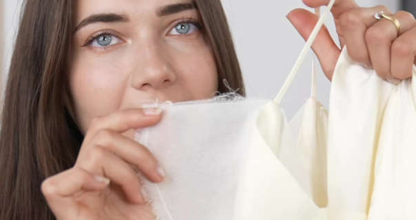 Roxxsaurus is seen in front of an off-white wall in her living room film set. She holds up a silk and chiffon ASOS dress. Roxi has her light brown hair loose and straight. She wears black mascara and soft foundation. Roxi is seen with her nails in a clear polish and with her diamond and gold wedding ring.