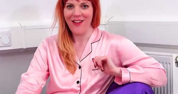 Kitty Quinzell sits in the corner of a white room with a full length wall power strip and a metal radiator behind her. Kitty has her long red hair loose and pushed back on one side. She appears in a chair covered in a purple bed sheet. Kitty wears a light pink black trim pyjama top with a red lipstick and black mascara.