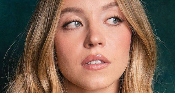 Sydney Sweeney is seen with her long blonde hair loose and in gentle waves. She wears a glossy dark pink lipstick with soft blue and black mascara. Sydney glances to one side. She stands in front of a dark green background for a photoshoot.