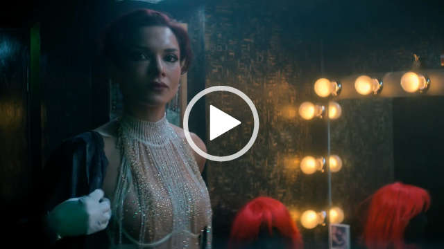 Amber Rose Revah stands next to a large mirror framed with lit yellow bulbs in a dark grey room. She wears a sheer glitter top with silver gloves and a robe. Amber has red tints in her short black hair.