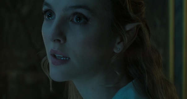Ann Skelly is seen in a large dark palace room. She wears her blonde hair down and pushed back and styled in tight curls over pointy ears. Ann stars in a dark pink matt lipstick and black mascara.