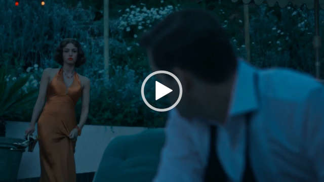 Mimi Keene wears a dark orange satin dress at dusk outdoors. She has her hair cut short and in tight curls. Mimi holds a pair of shiny heels in one had. She stands in front of a low cream wall and a border display of green plants and small flowers.