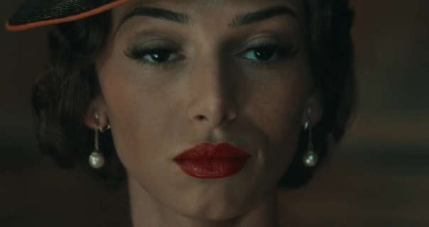 Mimi Keene stands in dock in a courtroom. She wears her hair in tight curls and cut short. Mimi is seen in a deep red velvet lipstick, curled eye lashes, a wide brim black and dark orange hat and a pair of drop pearl earrings.