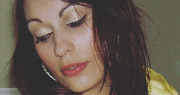 Vana Satin is seen with her black hair loose and in a messy style. She appears in front of a dark pea green wall and a white wood headboard. Vana wears a yellow nightie with gem encrusted hoop earrings, red blush, eyelash extensions and a pink gloss lipstick.