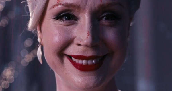 Gwendoline Christie smiles widely. Her silver blonde hair is tied back up in a bun. She wears a dark red matt lipstick and a pair of hanging cream seashell effect earrings. Gwendoline stands in front of sheer curtains and fairy lights at a dance. Her face has a few blood splatters across one eyebrow and down her nose.