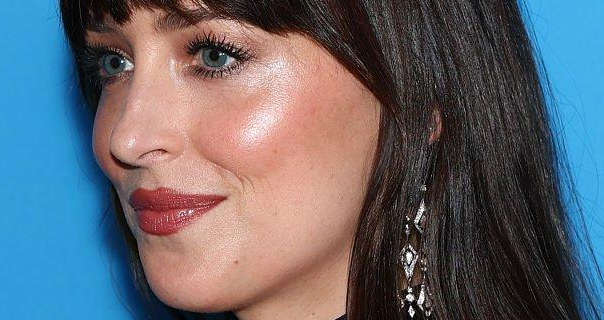 Dakota Johnson is seen from the side. She stands in front of a sky blue wall. Dakota wears a dark red lipstick with blush, black mascara and a pair of dangling silver and gemstone earrings. Her long black hair is loose, straight and down.