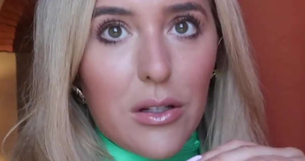 Em Sheldon appears in a hotel room in Tuscany. She is seen in front of a terracotta wall. Em has her long thick blonde hair straight and down. She wears a green dress with a pair of gold earrings, mascara and a glossy pink lipstick.