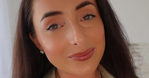 Hazel Maria Wood smiles and looks into the camera. She stands in front of a dark cream wall in her home living room. Hazel wears her thick hair loose and straightened. She appears in a pair of emerald style stud earrings and a matching gemstone necklace from Italo Jewelry.