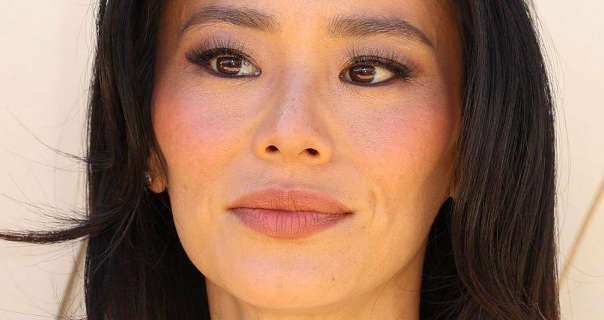 Jamie Chung is seen with her black hair long, straight and loose. She wears blush with a pair of small earrings, a matt pink lipstick, black eyeliner and mascara. She stands in front of a pale gold wall and looks to the side.