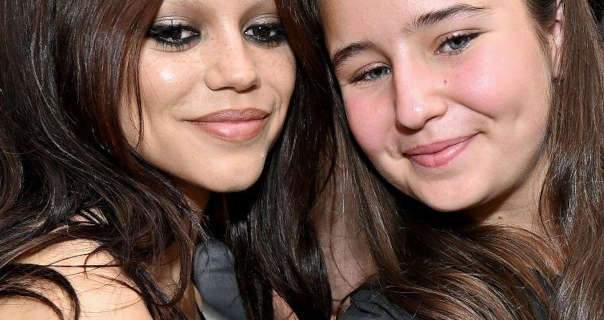 Jenna Ortega takes a selfie with a fan outdoors at an event. Jenna wears her long black hair loose and slightly curled. She appears in a pale pink matt lipstick, black eyeliner and black mascara.