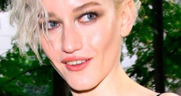 Julia Garner walks past a few small leafy green trees. She wears her white blonde hair loose and cut short. Julia appears in mascara, dark red eye shadow, pink lipstick and a little red blush.