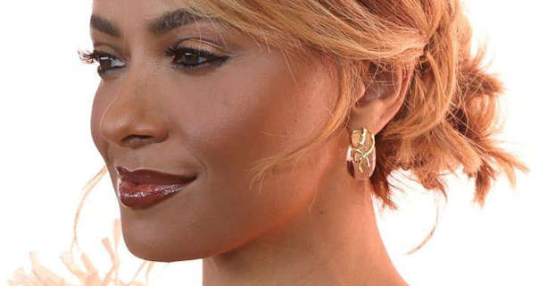 Kat Graham wears her hair tied back and in a reddish blonde colour. She appears in a dark red gloss lipstick, mascara and a pair of large gold earrings. Kat stands in front of a bright white wall in a tassel jacket.