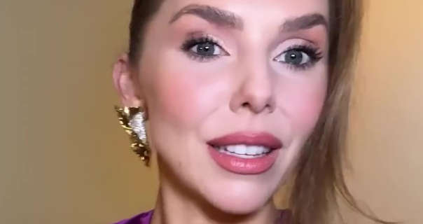 Laura Blair is seen in a dark pink gloss lipstick, blush, mascara and a pair of large gold sheet earrings. She wears her blonde hair tied up with a section pulled down to frame one side of her face. She stands in front of a cream hotel room wall in Japan.