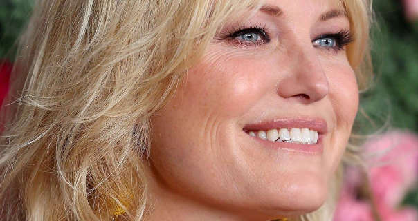 Malin Akerman is seen from a side angle. She smiles with her blonde hair loose and frizzy. Malin stands in front of a green leaf and pink rose wall. She wears black mascara with soft blush, light foundation and a dark pink lipstick.